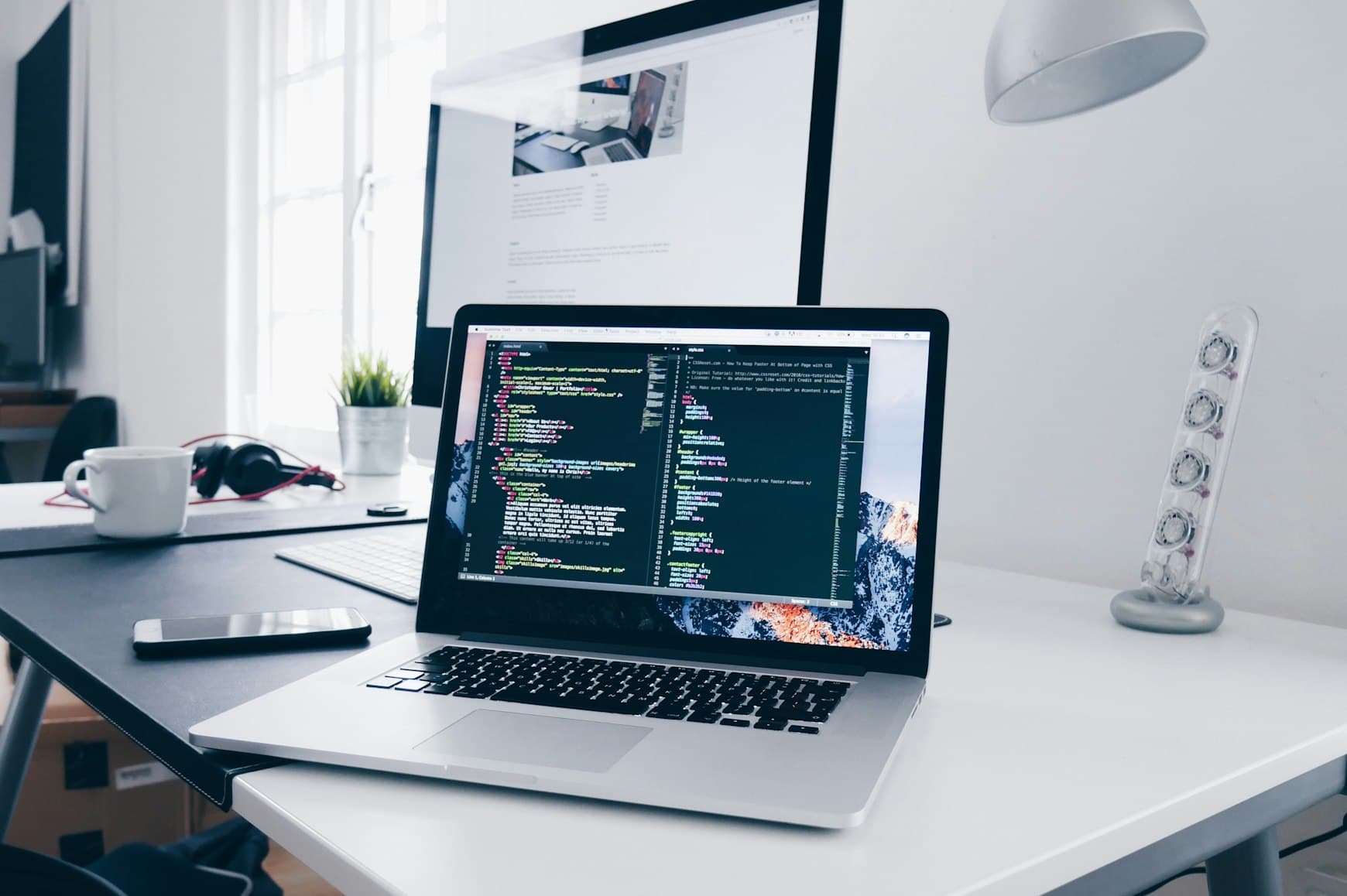 A laptop on a white desk showing a code editor, with a second monitor behind it